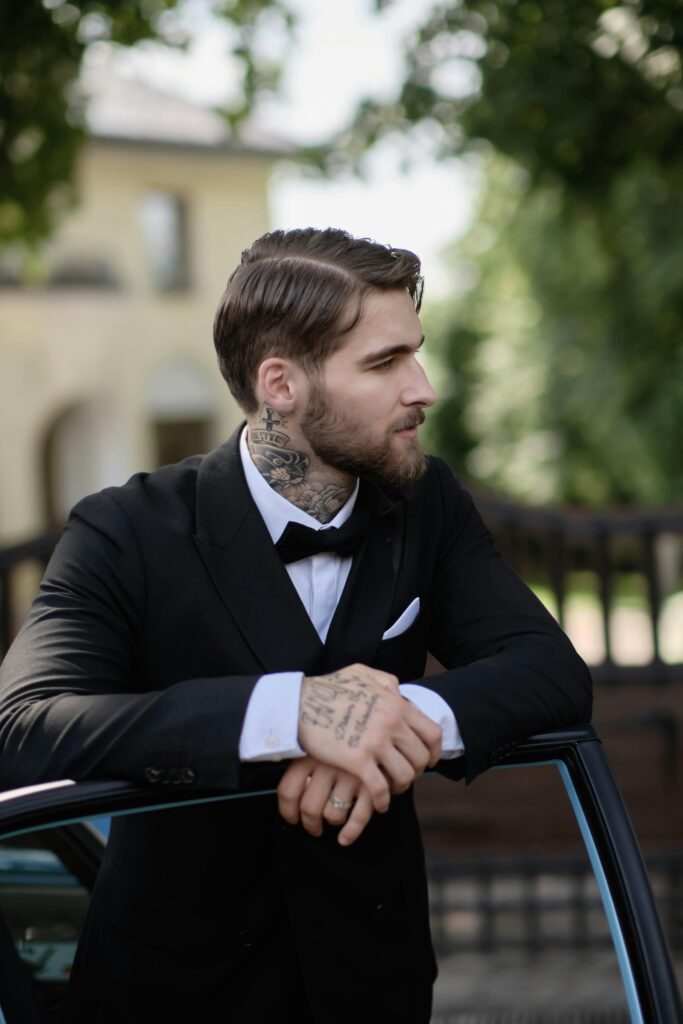Elegantly dressed man with tattoos leans on a car door in an outdoor setting.